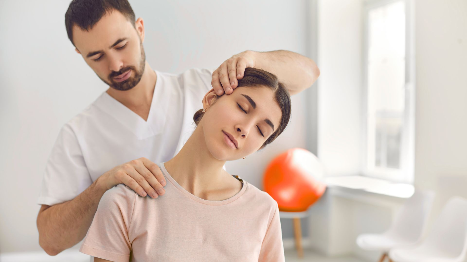 Healthcare professional in white uniform performing neck stretch on seated patient in pink shirt, eyes closed in relaxation.