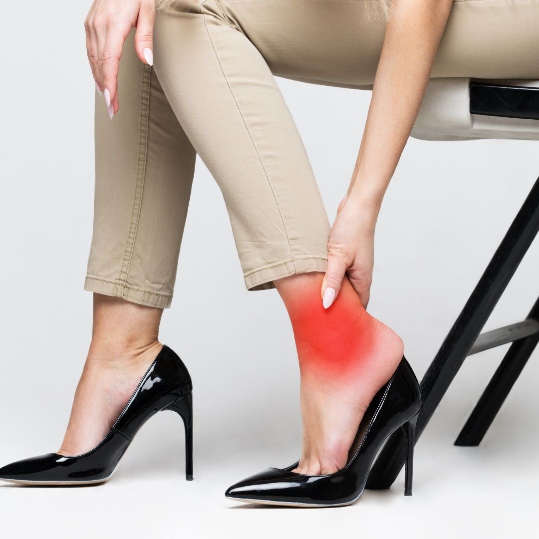Foot pain from high heels—woman seated, clutching red, irritated ankle in beige pants and black stilettos.