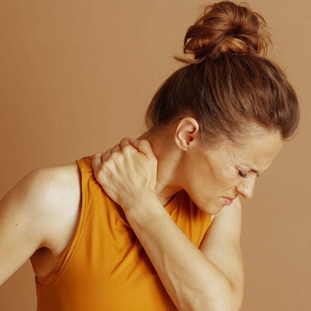Person holding left side of neck in pain, wearing orange top against plain background.