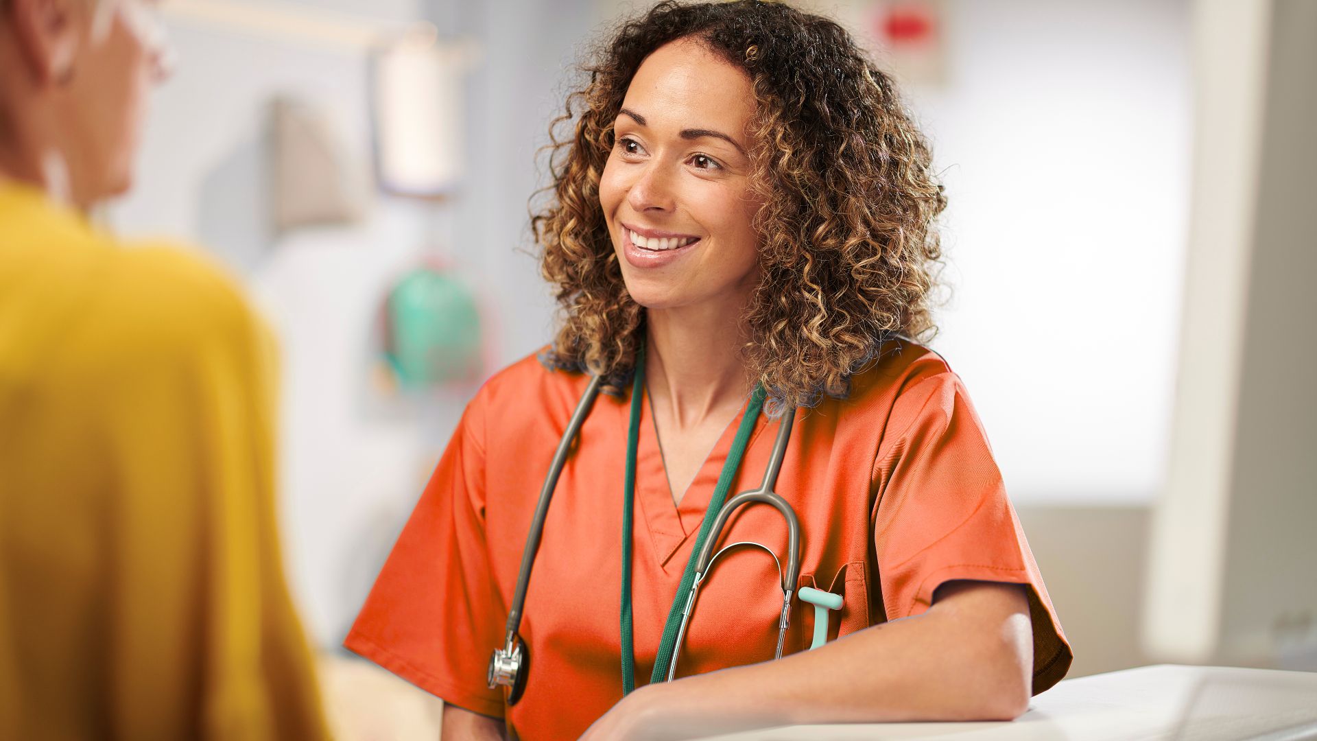 Healthcare professional in orange scrubs with stethoscope speaking to a patient in a clinical setting.