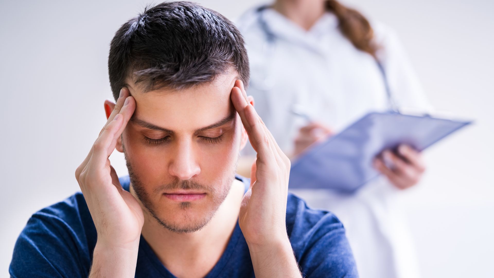 Person holding temples in discomfort while clinician observes with clipboard in clinical setting.