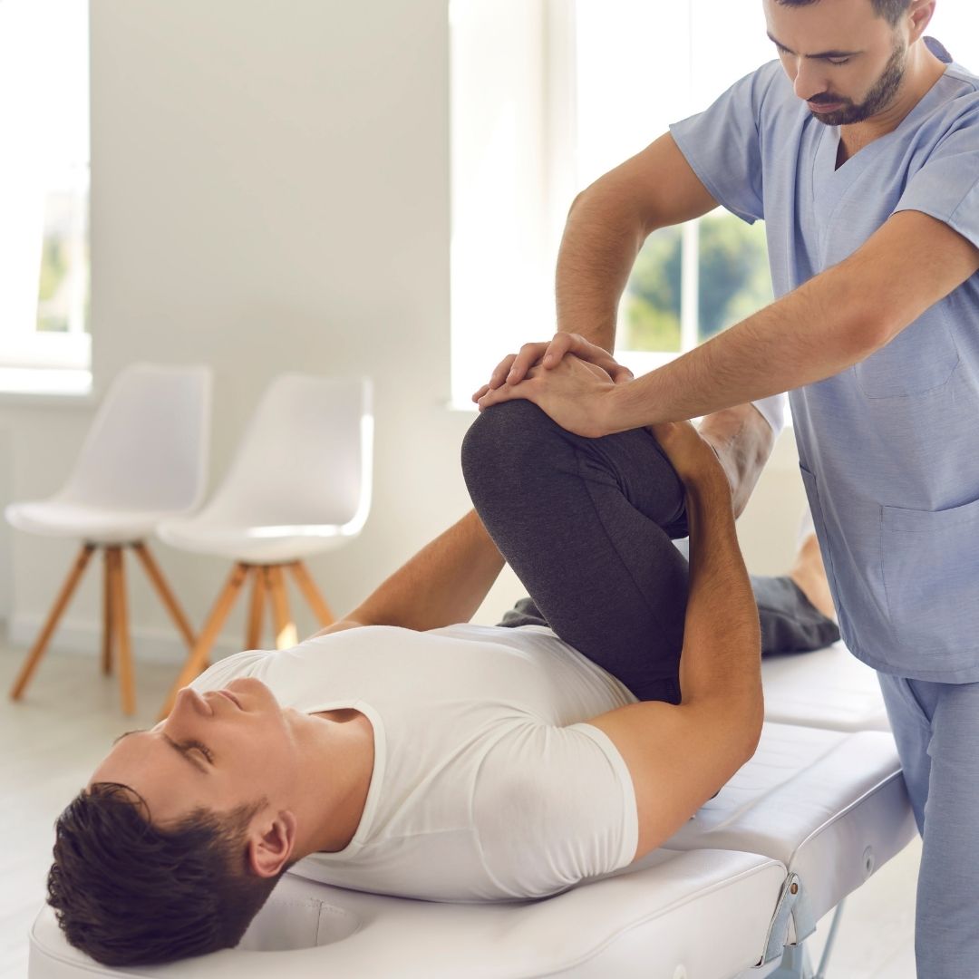 Physical therapist performing hip stretch on patient lying on treatment table in a bright clinical setting.