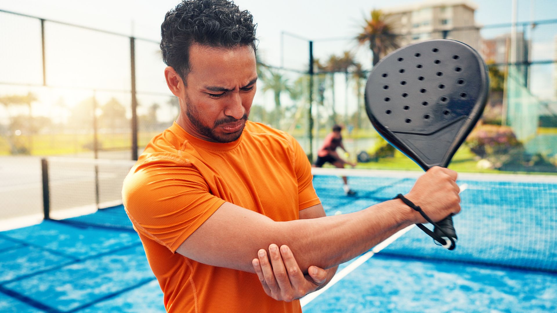 Padel player gripping elbow in pain during outdoor match, suggesting possible sports-related injury.