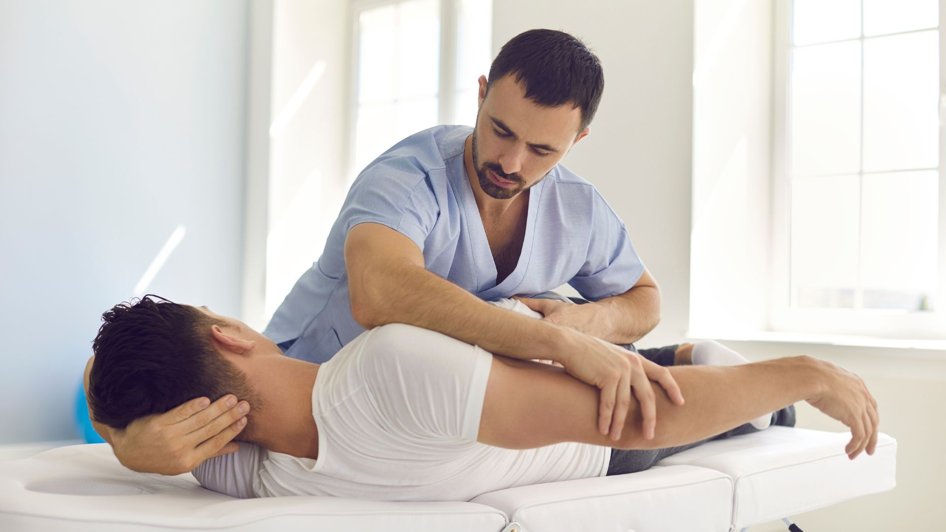 A physiotherapist in a blue uniform adjusts a patient's shoulder during a session. The patient lies on a white bed in a bright room, conveying care and focus.